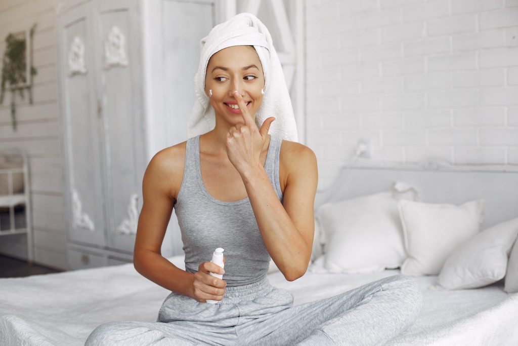 woman sat on bed with hair in a towel putting on skin care