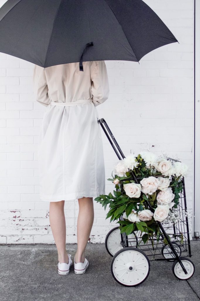 Woman wearing beige trench coat facing wall holding umbrella next to a trolley of white flowers